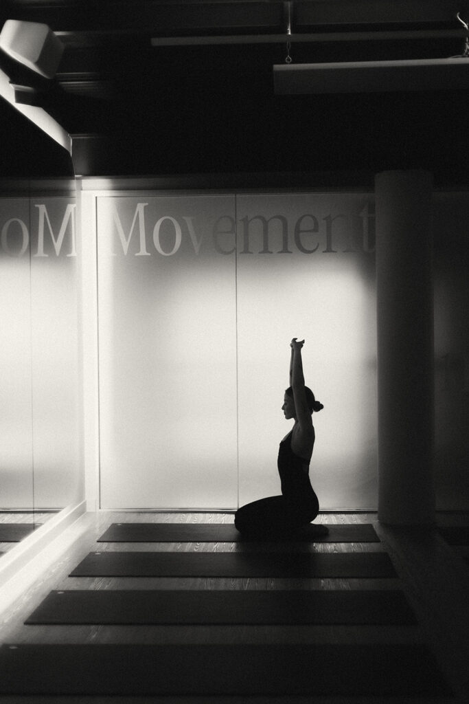 Silhouette of a woman seated in a yoga pose with arms raised, captured in a minimal movement studio by Sunshine Coast wellness photographer.