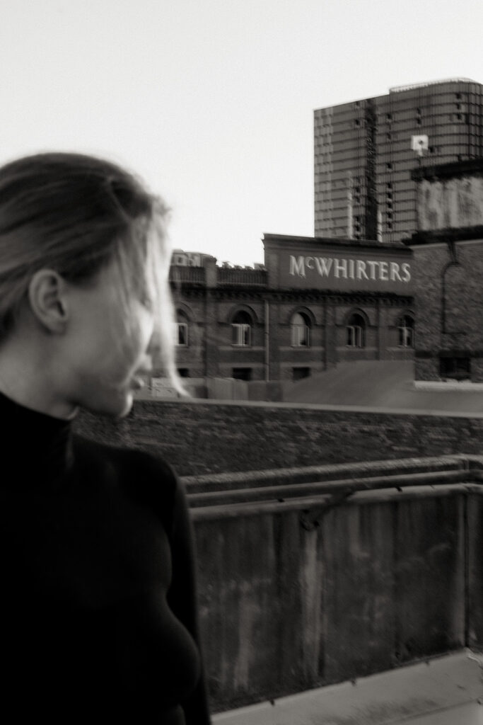Black-and-white image of a woman in a turtleneck on a rooftop, blurred in the foreground with the McWhirters building in focus behind her, photographed by Sunshine Coast brand photographer.