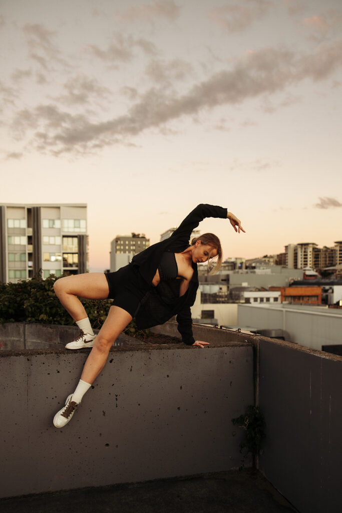 Woman in athletic wear holding a strong, expressive movement pose on a rooftop ledge at sunset, captured by Sunshine Coast movement and brand photographer.