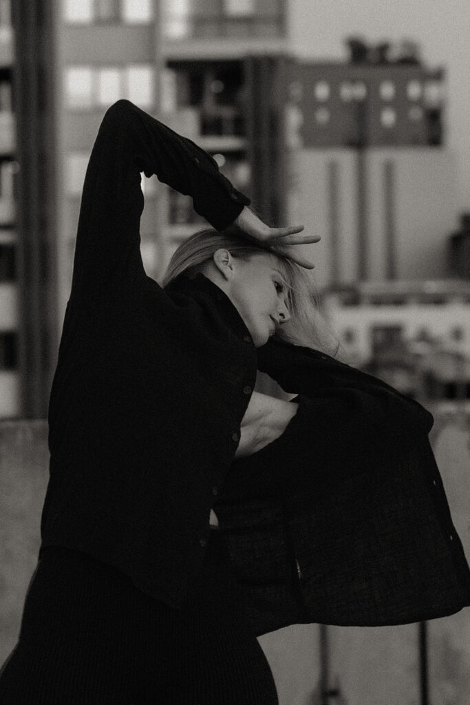 Black-and-white photo of a woman moving expressively with arms raised, set against a blurred city skyline in Brisbane