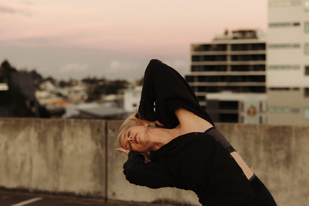 Woman arching into a fluid, expressive pose on a rooftop at sunset, wearing black and framed against soft pastel skies and city buildings, captured by Sunshine Coast movement photographer.