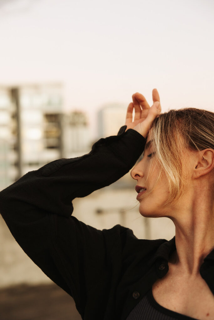 Close-up of a woman with eyes closed and hand raised to her forehead, bathed in soft golden light on a rooftop, photographed by Sunshine Coast brand photographer.