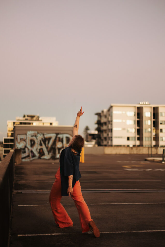 Woman in wide-legged pants reaching toward the sky in an expressive pose on a rooftop car park, with graffiti and city buildings in the background, captured by Sunshine Coast movement photographer.