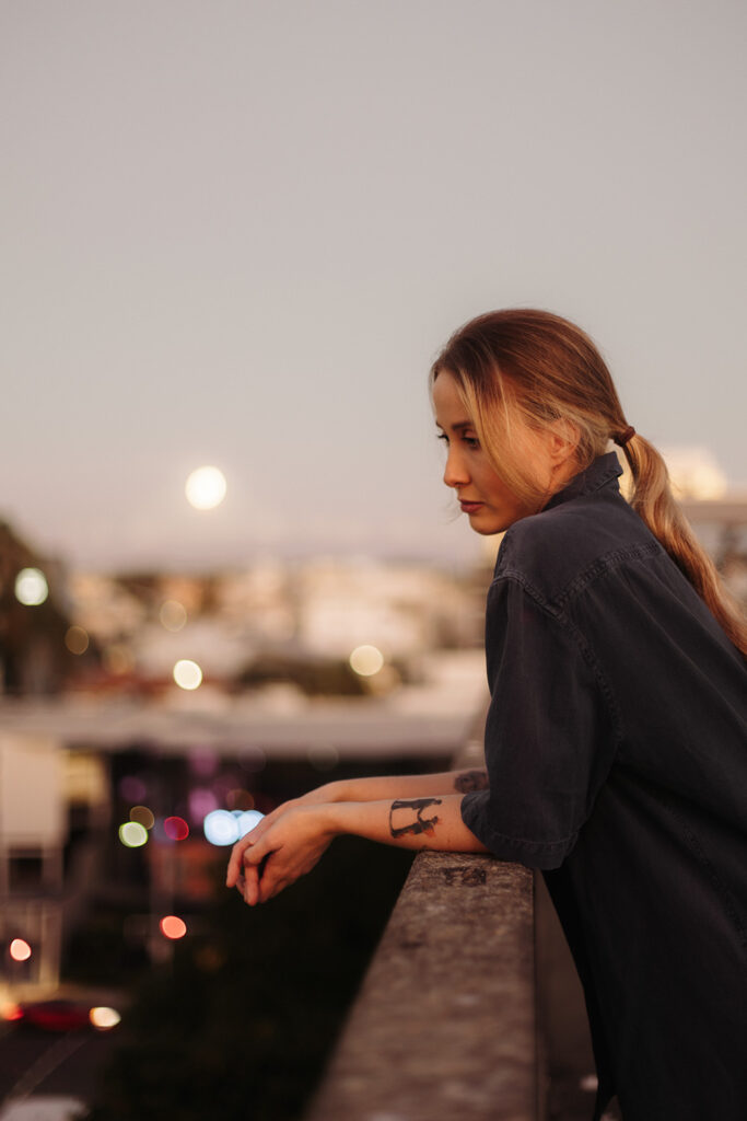 Woman leaning on a rooftop ledge at dusk, looking out over a blurred city with a full moon in the background, captured by Sunshine Coast brand photographer.