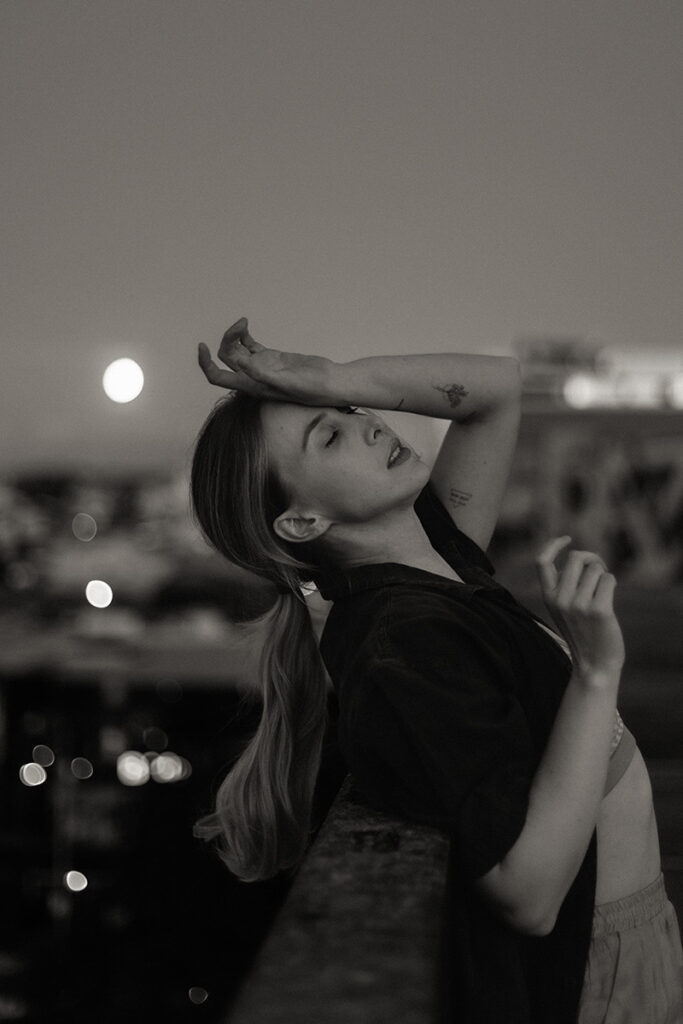 Black-and-white image of a woman leaning on a rooftop ledge under the full moon, eyes closed in a moment of reflection, captured by a Sunshine Coast brand photographer.