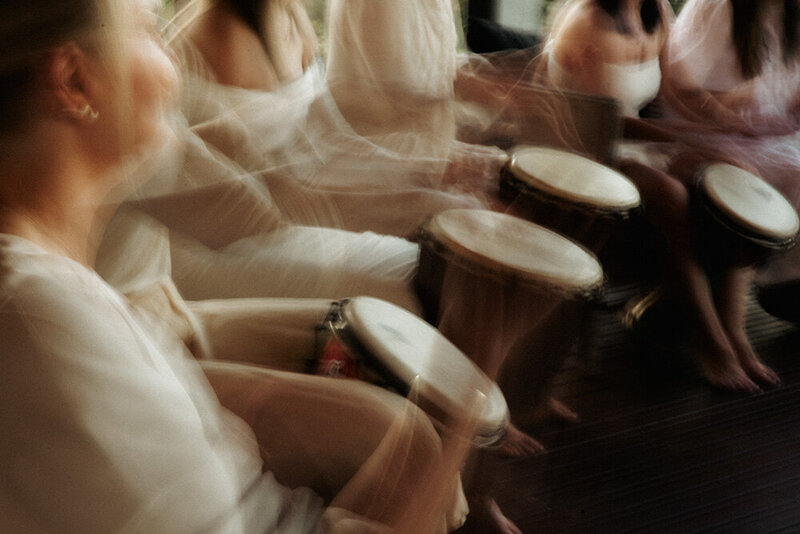 A group of women in soft, neutral-toned clothing playing hand drums in a blurred motion, creating a sense of rhythm and connection.