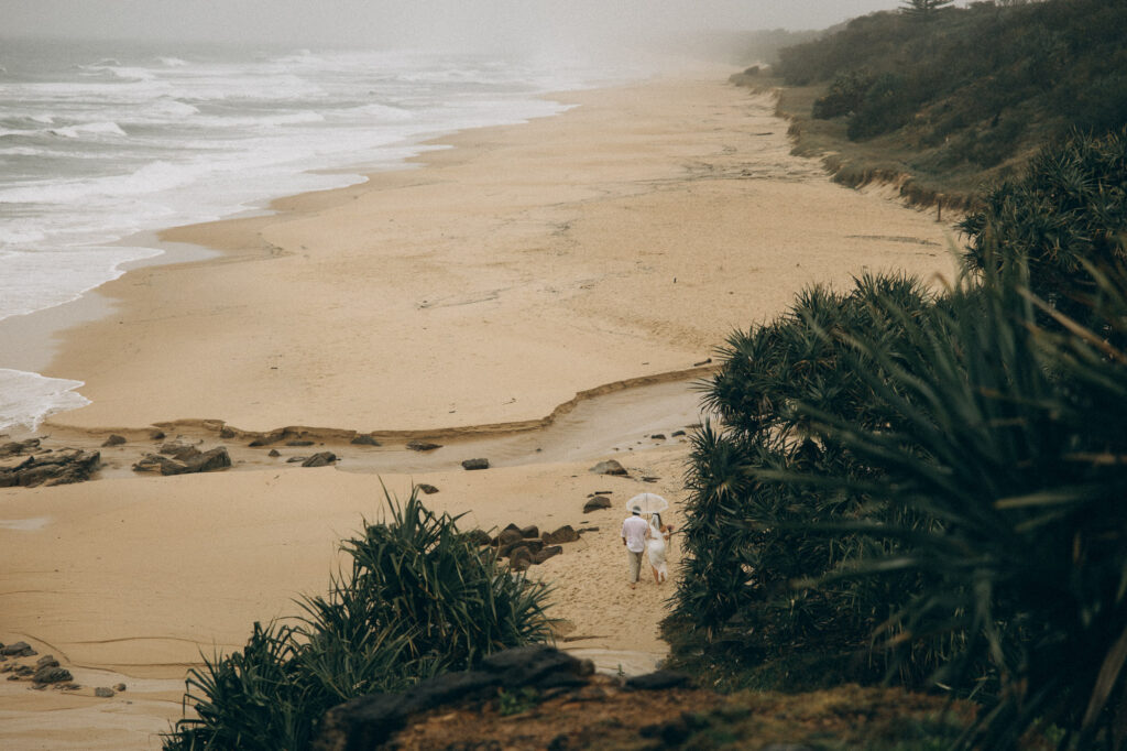 Bride and groom walking under umbrella along deserted Coolum Beach during rainy Sunshine Coast elopement