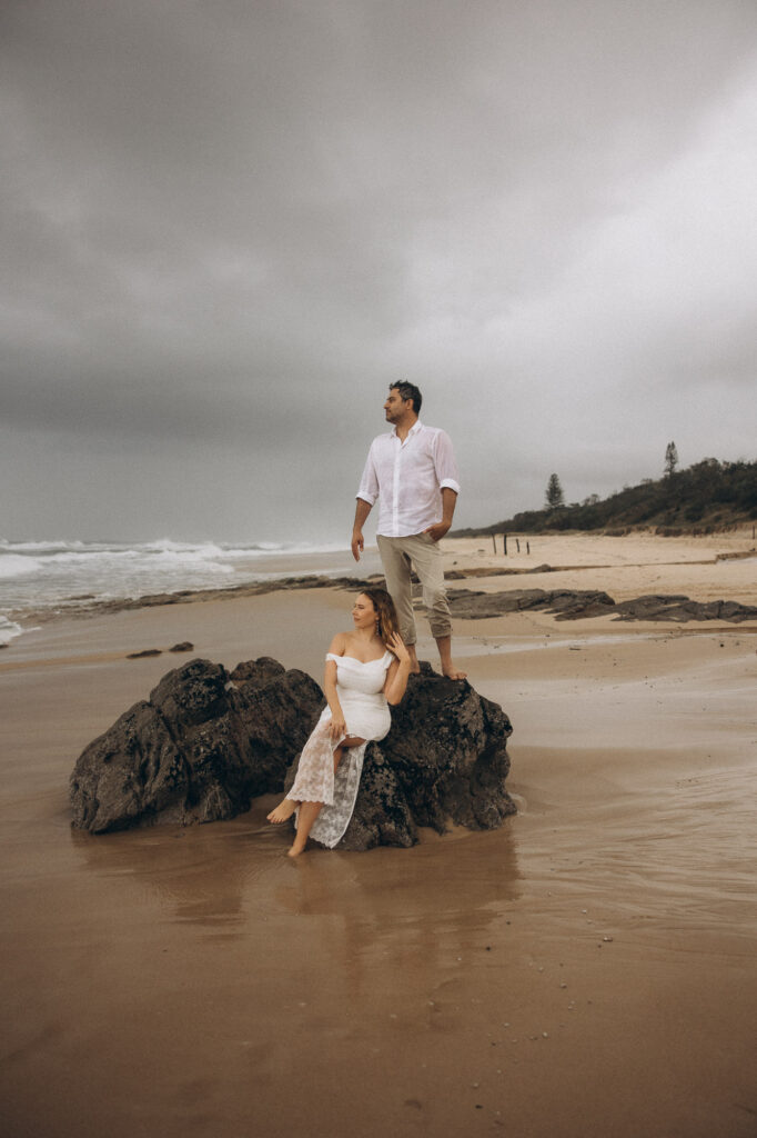 Couple standing on rocks during moody Coolum Beach elopement, Sunshine Coast wedding photography capturing cinematic coastal love story