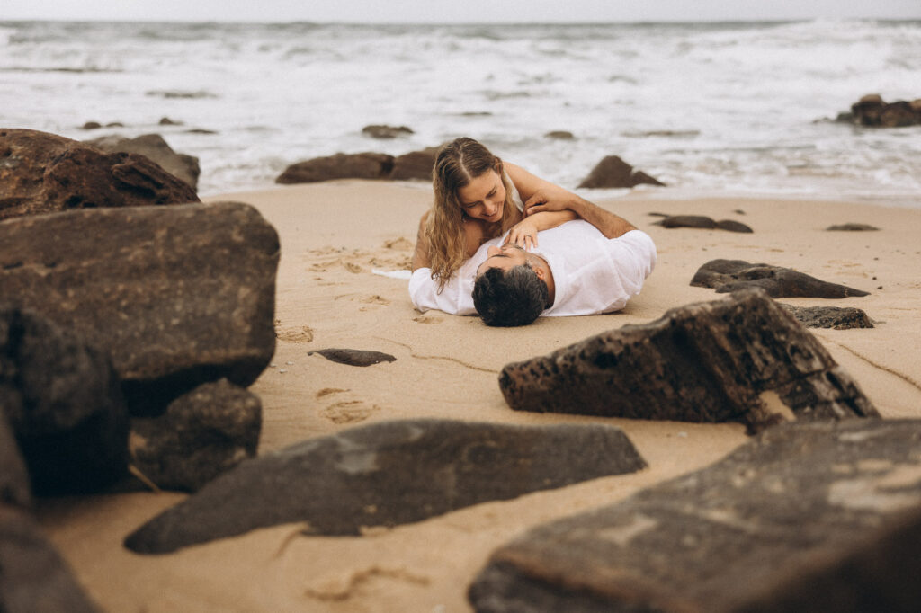 Playful bride and groom lying on sand between rocks during intimate Sunshine Coast beach elopement