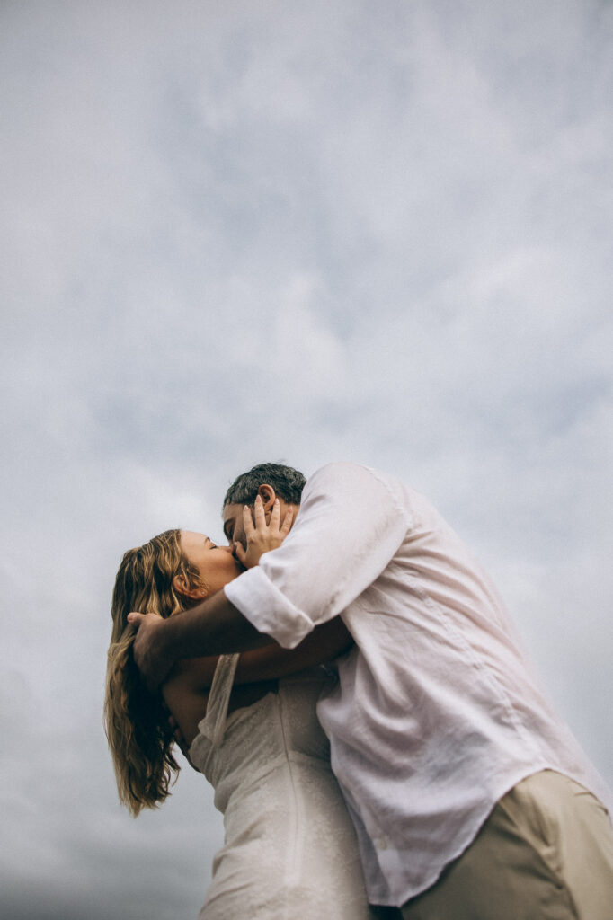 Bride and groom kissing beneath cloudy sky during cinematic Sunshine Coast elopement at Coolum Beach