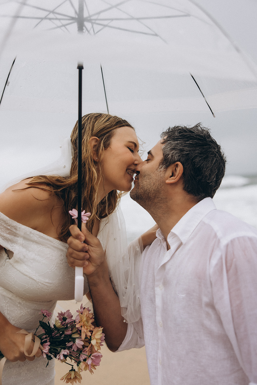 Bride and groom share a kiss under a clear umbrella during their rainy Coolum Beach elopement on the Sunshine Coast, captured by Nicci K Photography.