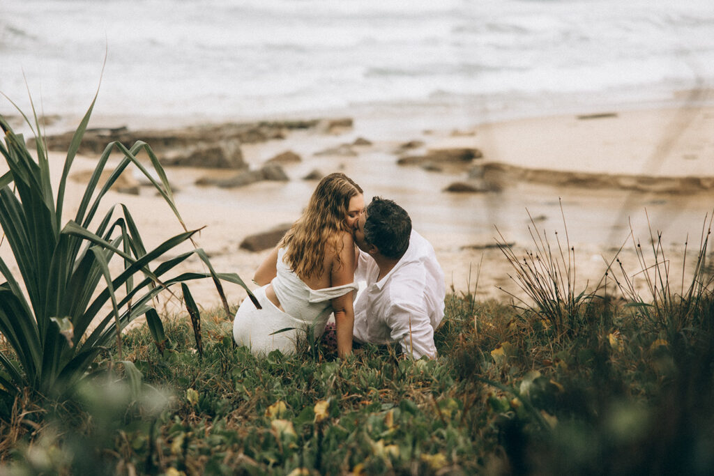 Couple kissing on grassy dunes overlooking Coolum Beach during intimate Sunshine Coast elopement, captured by Nicci K Photography