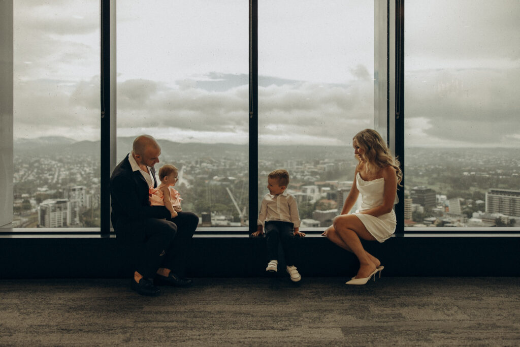 Modern Brisbane Registry Office elopement with city skyline views. Bride in a chic short white dress and groom in a tailored black suit sit by the floor-to-ceiling windows with their two young children, creating a warm, intimate family moment. Captured by Nicci K Photography — cinematic Brisbane elopement photographer specialising in emotive, storytelling imagery for modern couples and families.