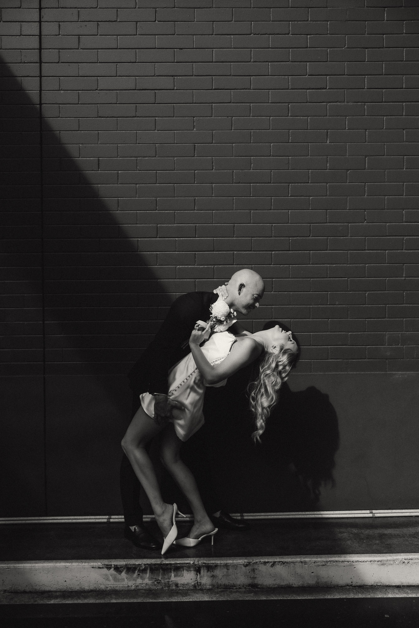 Cinematic black and white photo of a stylish couple during their Brisbane City elopement, sharing an intimate dip kiss against a brick wall with dramatic light and shadow. Captured by Nicci K Photography, Brisbane elopement photographer known for emotive, cinematic storytelling.