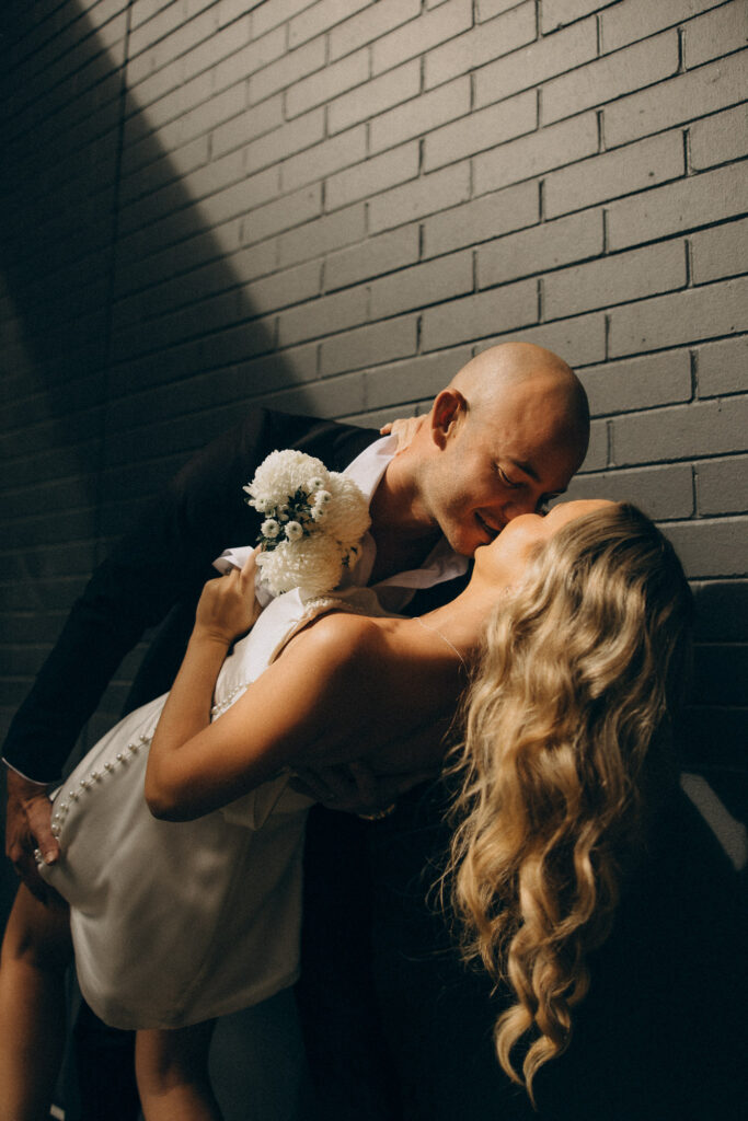 Modern Brisbane City elopement captured in cinematic light — groom dips bride for a romantic kiss against an urban brick wall. Bride wears a short pearl-detailed dress holding white florals. Shot by Nicci K Photography, Brisbane elopement photographer creating cinematic, emotive love stories for modern couples.