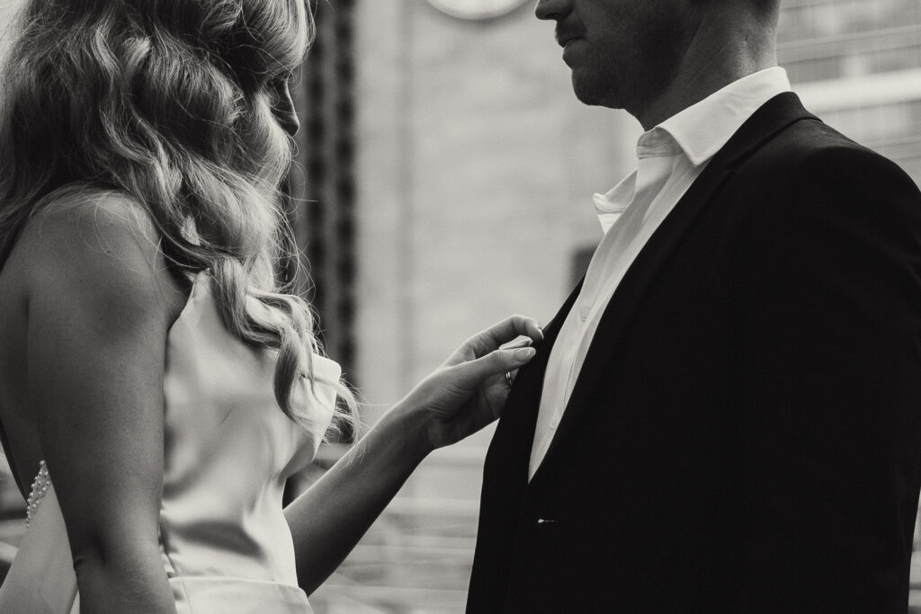 Cinematic close-up black and white photo of an intimate moment between a couple during their Brisbane City elopement. The bride gently adjusts the groom’s jacket, capturing a quiet, emotional connection amid urban architecture. Shot by Nicci K Photography, Brisbane elopement photographer known for raw, emotive storytelling.