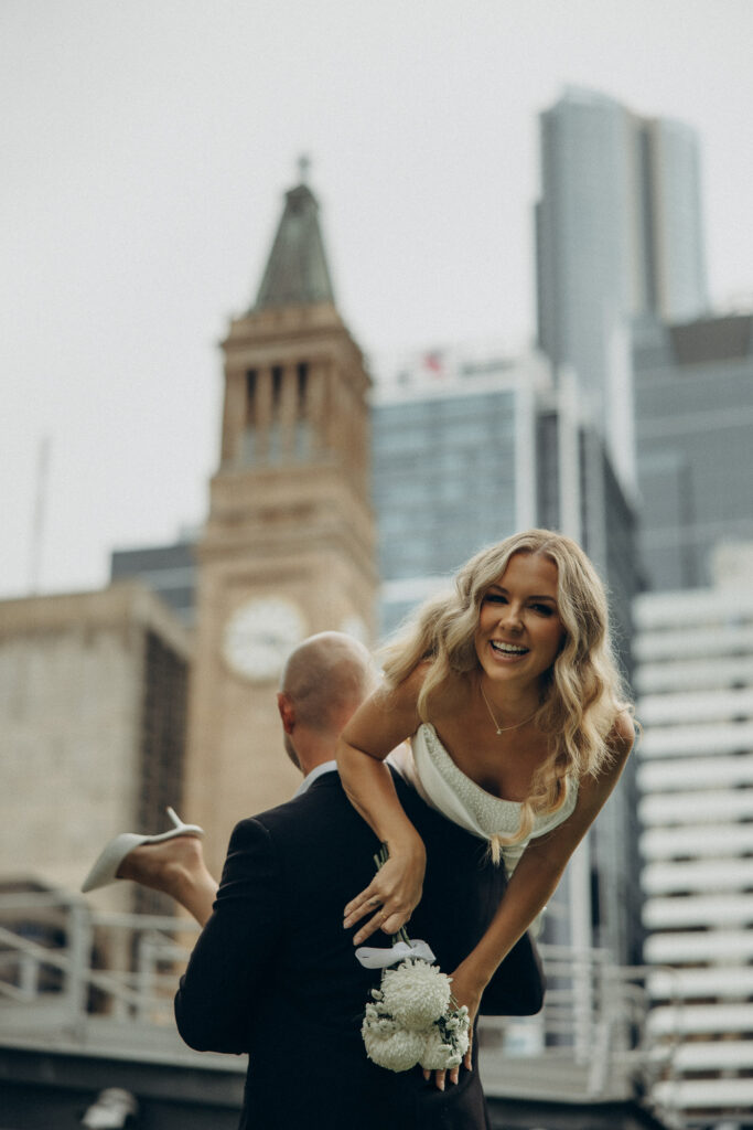 Playful Brisbane City elopement moment captured near King George Square and City Hall clock tower. Bride laughs joyfully while being carried by the groom, bouquet in hand, surrounded by Brisbane’s skyline. Captured by Nicci K Photography — cinematic, emotive Brisbane elopement and wedding photographer for modern couples.