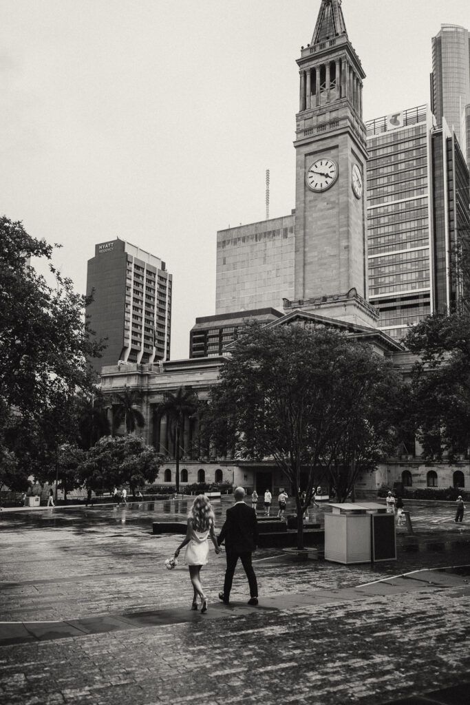 Cinematic black and white photo of a couple walking hand in hand after their Brisbane City elopement near the iconic Brisbane City Hall clock tower. Captured by Nicci K Photography, Brisbane elopement and wedding photographer specialising in cinematic, emotive storytelling for modern couples.