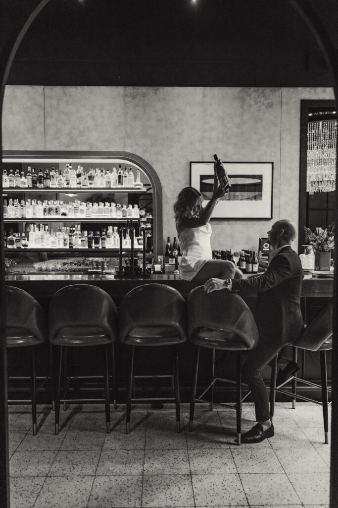 Cinematic black and white photo of a Brisbane elopement celebration inside Rothwell’s Bar. Bride sits on the bar counter holding a champagne bottle while the groom smiles beside her. Captured by Nicci K Photography — Brisbane elopement photographer documenting modern, intimate, and romantic city weddings.