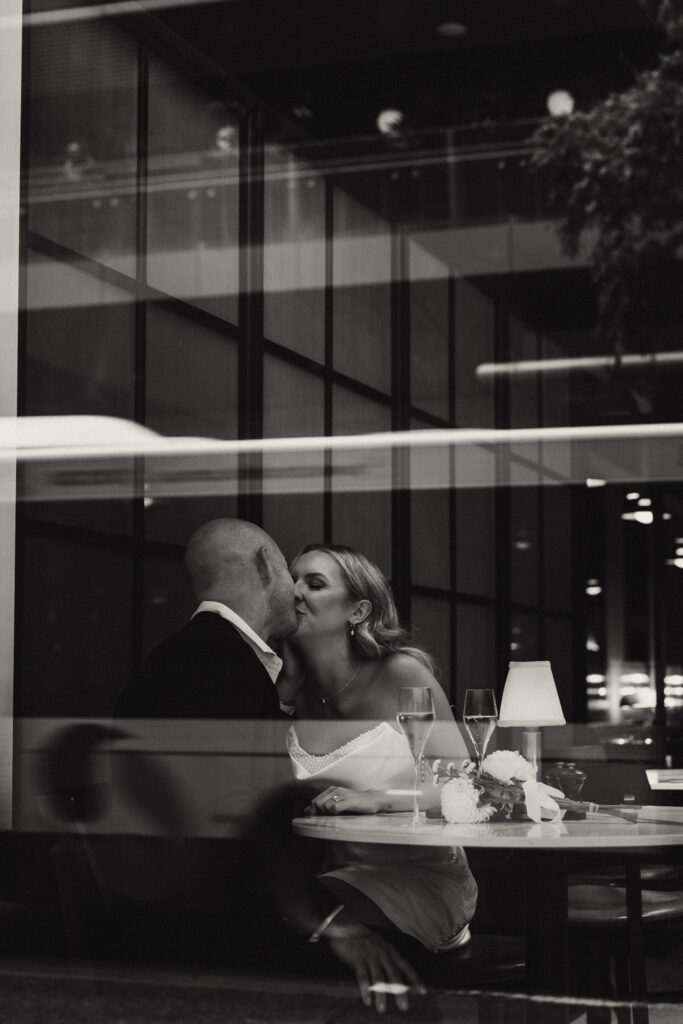 Romantic Brisbane elopement moment captured through the window of a city restaurant. Bride and groom share an intimate kiss over champagne, reflecting the elegance of an urban Brisbane registry wedding. Captured by Nicci K Photography — cinematic and emotive Brisbane elopement photographer.
