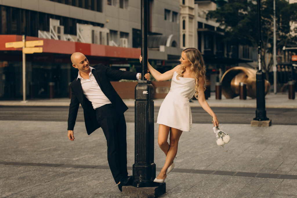 Playful Brisbane City elopement photo of a modern couple swinging around a street lamp in golden afternoon light. The bride holds her bouquet, laughing as the groom leans toward her in a classic black suit. Captured by Nicci K Photography — cinematic Brisbane elopement photographer specialising in authentic, storytelling imagery for modern couples.