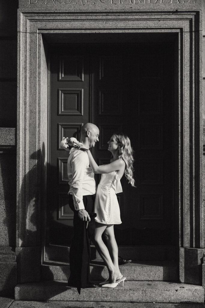 Cinematic black and white Brisbane elopement photo captured on the marble steps of Treasury Brisbane. Bride in a short satin dress stands elegantly while the groom sits below, jacket over shoulder, creating a timeless, editorial-style wedding portrait. Captured by Nicci K Photography — Brisbane elopement photographer for modern, cinematic love stories.
