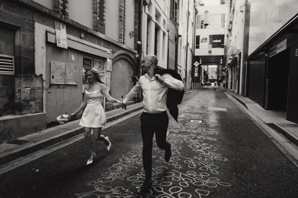 Candid black and white Brisbane City elopement moment of a couple running hand in hand through an urban alleyway. The bride laughs joyfully, holding her bouquet, as the groom carries his jacket. Captured by Nicci K Photography — Brisbane elopement and wedding photographer creating cinematic, emotive storytelling imagery.