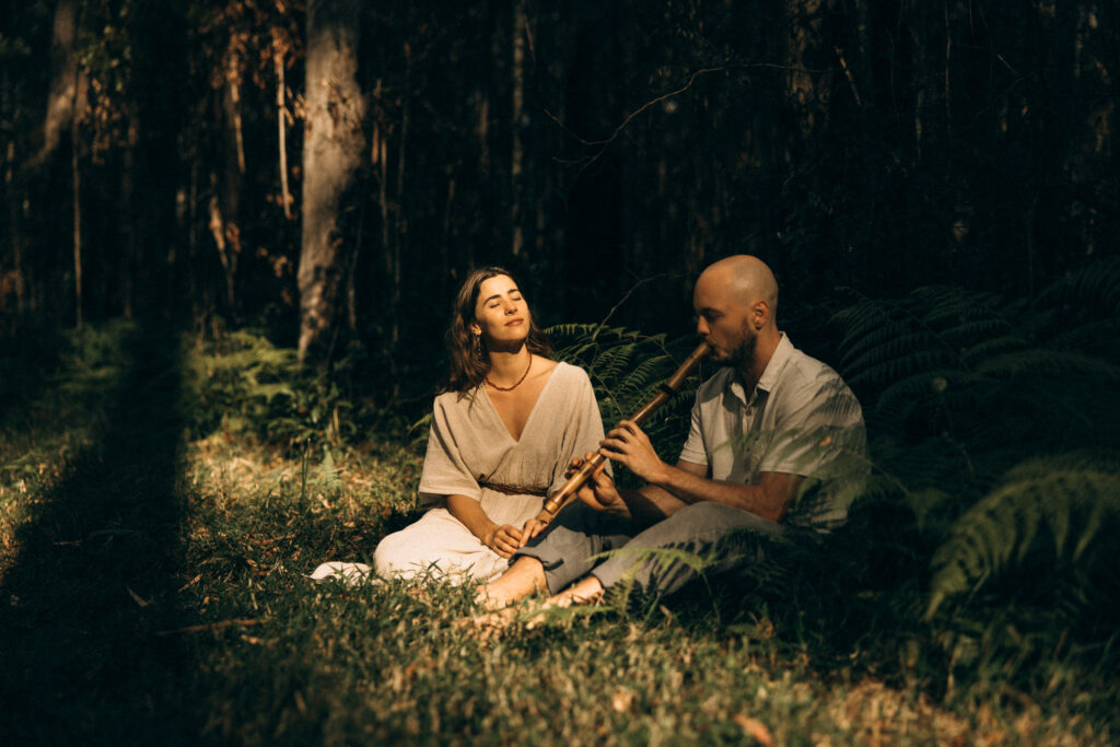 Couple sitting barefoot in the Maleny rainforest during a cinematic Sunshine Coast storytelling session, with the man playing a wooden flute while his partner listens in the golden light. A romantic forest couples shoot inspired by van life, connection, and slow living in the Sunshine Coast Hinterland.