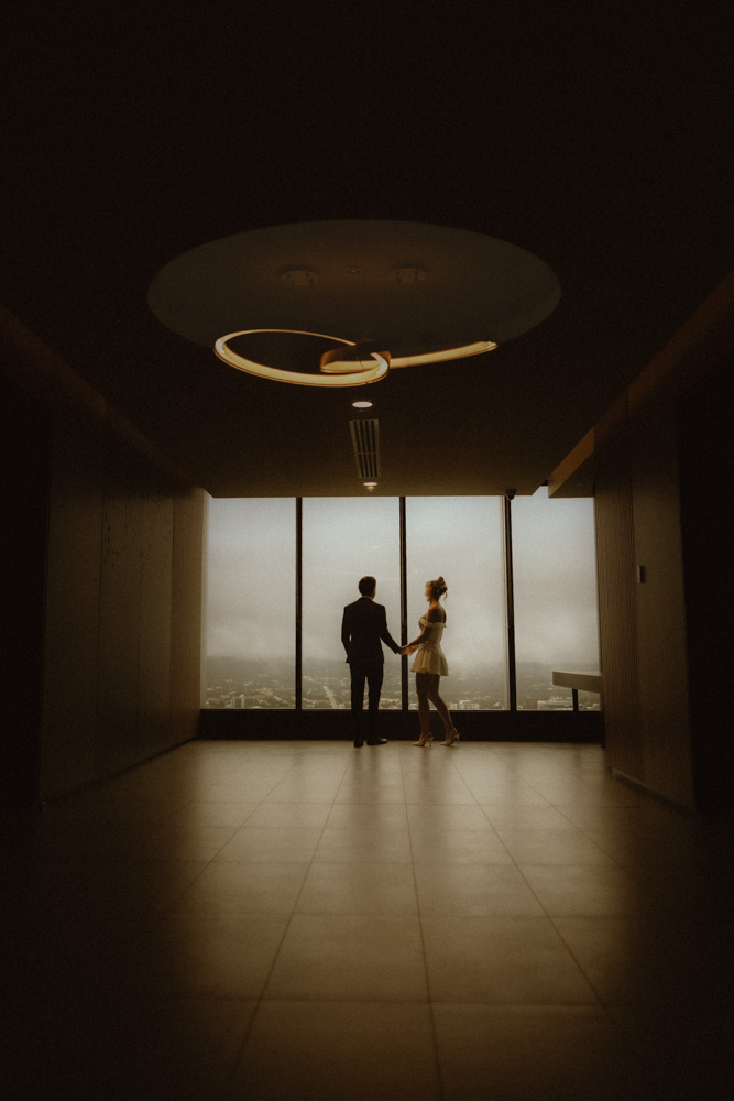 Bride and groom walking hand in hand through a dim hallway toward a lit window after their Brisbane Registry Office ceremony