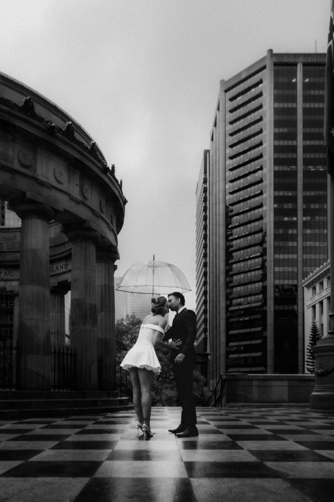 Black and white wide shot of bride and groom under an umbrella at Anzac Square's sandstone colonnade