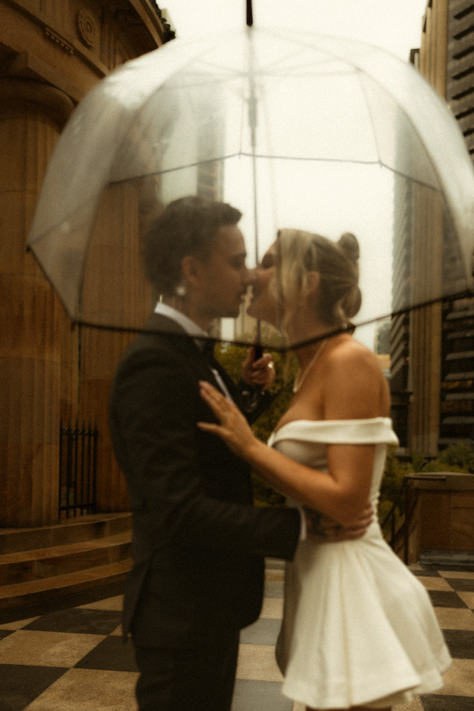 Sepia shot of bride and groom under umbrellas beside the Anzac Square colonnade