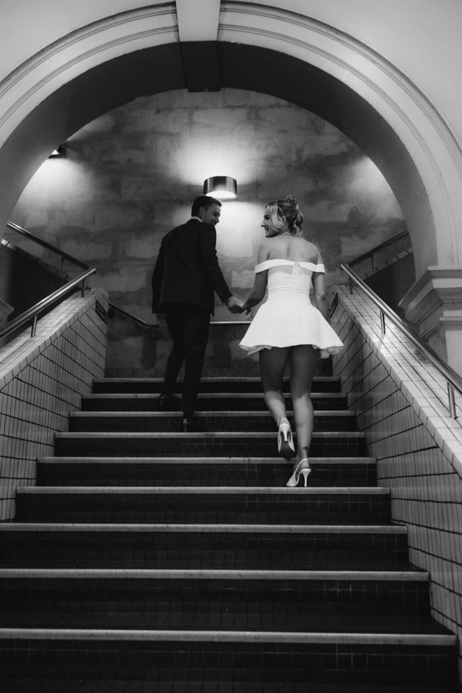 Black and white wide shot of bride and groom descending a station stairwell hand in hand
