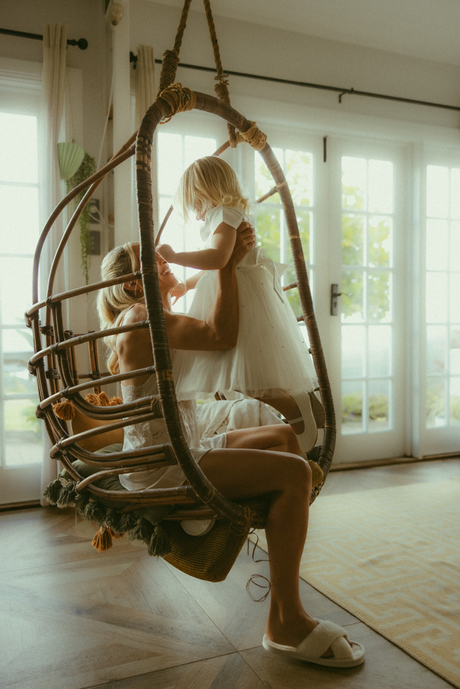 bride holding her daughter wearing flower girl dress in a egg chair in the bridal suite
