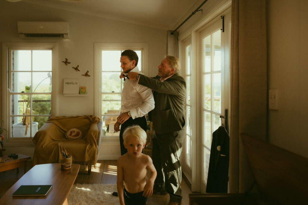Brides father helping him into tuxedo near a window in the grooms suite before their elopement