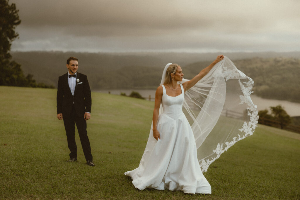 Editorial pose of bride waving her veil on the grass with Sunshine Coast hinterland in background as groom watched