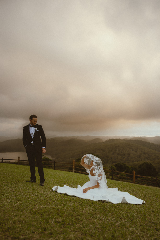Editorial pose of bride sitting on the grass with veil over her head while the groom is standing back admiring her 